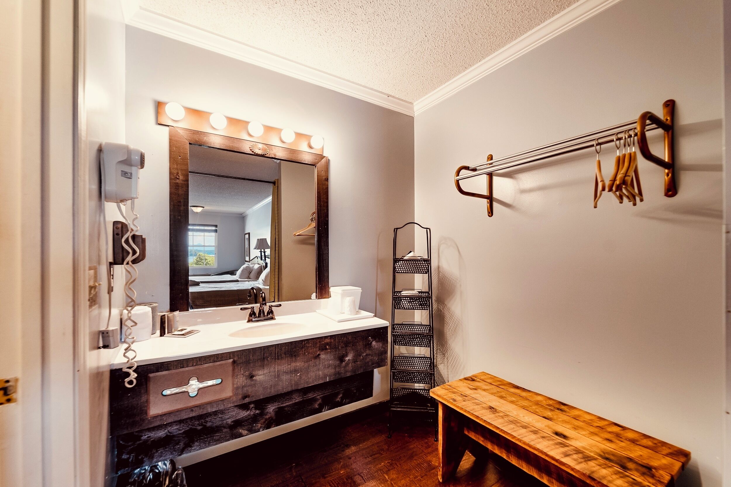 A bathroom with a vanity, a mirror with lights, a towel rack, a bench, and a shelf.