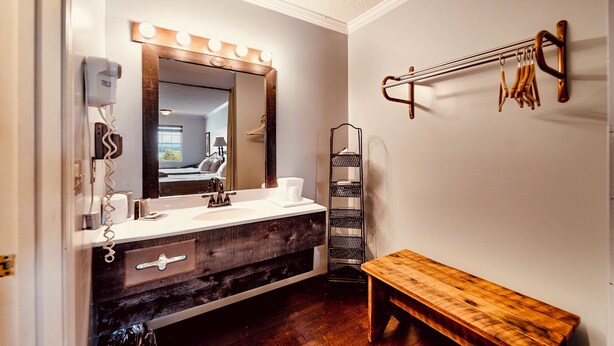 A bathroom with a vanity, a mirror with lights, a towel rack, a bench, and a shelf.