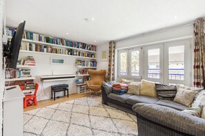 A living room with a sofa, a television, a bookshelf, and a window with curtains. - GuestReady - Charming London Flat with Balcony (London)