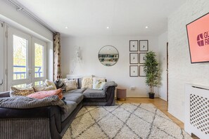 A living room with a sectional sofa, a round mirror, framed pictures, a potted plant, and a white brick wall. - GuestReady - Charming London Flat with Balcony (London)