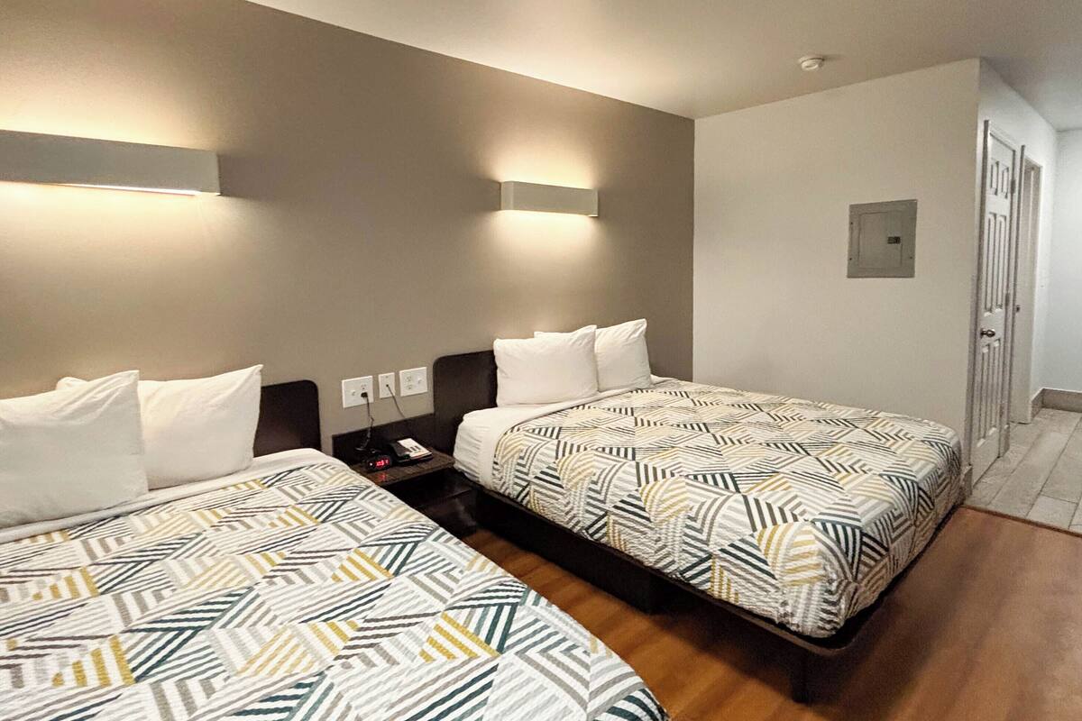 Habitación de hotel con dos camas, una mesita de noche, una lámpara en la pared y una puerta a la derecha.