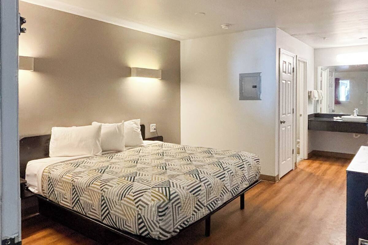 Un dormitorio con colcha estampada, una mesita de noche, una lámpara de pared, una puerta y un baño con lavabo y espejo.