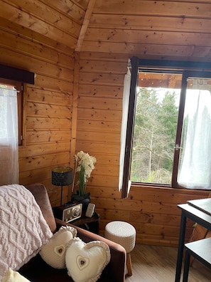 Interior acogedor de una cabaña con paredes de madera, una cama con manta blanca, una mesita y una ventana con vista a árboles.