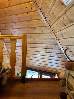 Un dormitorio de madera en el ático, con techo inclinado, estructura de vigas de madera y una ventana pequeña.