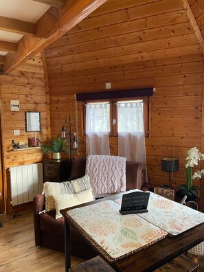 L’intérieur d’un chalet accueillant, avec un plafond en bois, une table à manger sur laquelle se trouve un ordinateur portable, un canapé, un radiateur et une fenêtre avec des rideaux.