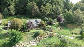 Une maison entourée d’un jardin luxuriant, avec un mur en pierre, une terrasse en bois et une vue sur une colline boisée.