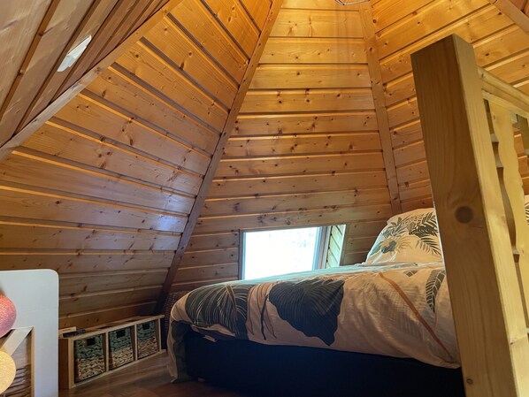 Une pièce de style cabane en bois, avec un plafond en pente, un lit recouvert d’une literie à motifs et une fenêtre laissant entrer la lumière naturelle.
