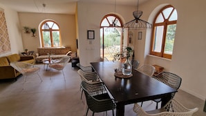 A dining area with a table, chairs, and a sofa. There are large windows with arched tops, a hanging light fixture, and a potted plant on the table. - Beautiful vacation villa in the heart of Puglia Castellana Grotte (Puglia)