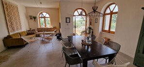 A dining area with a table, chairs, and a sofa. There are large windows with arched tops, a hanging light fixture, and a potted plant on the table. - Beautiful vacation villa in the heart of Puglia Castellana Grotte (Puglia)