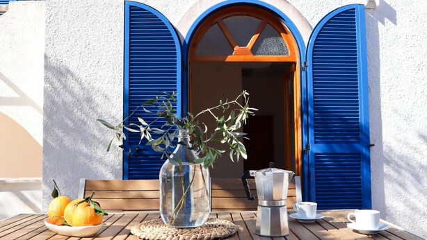 A table with a clear glass vase, a plate of lemons, a coffee maker, and cups on a wooden table outside a building with blue shutters. - Beautiful vacation villa in the heart of Puglia Castellana Grotte (Puglia)