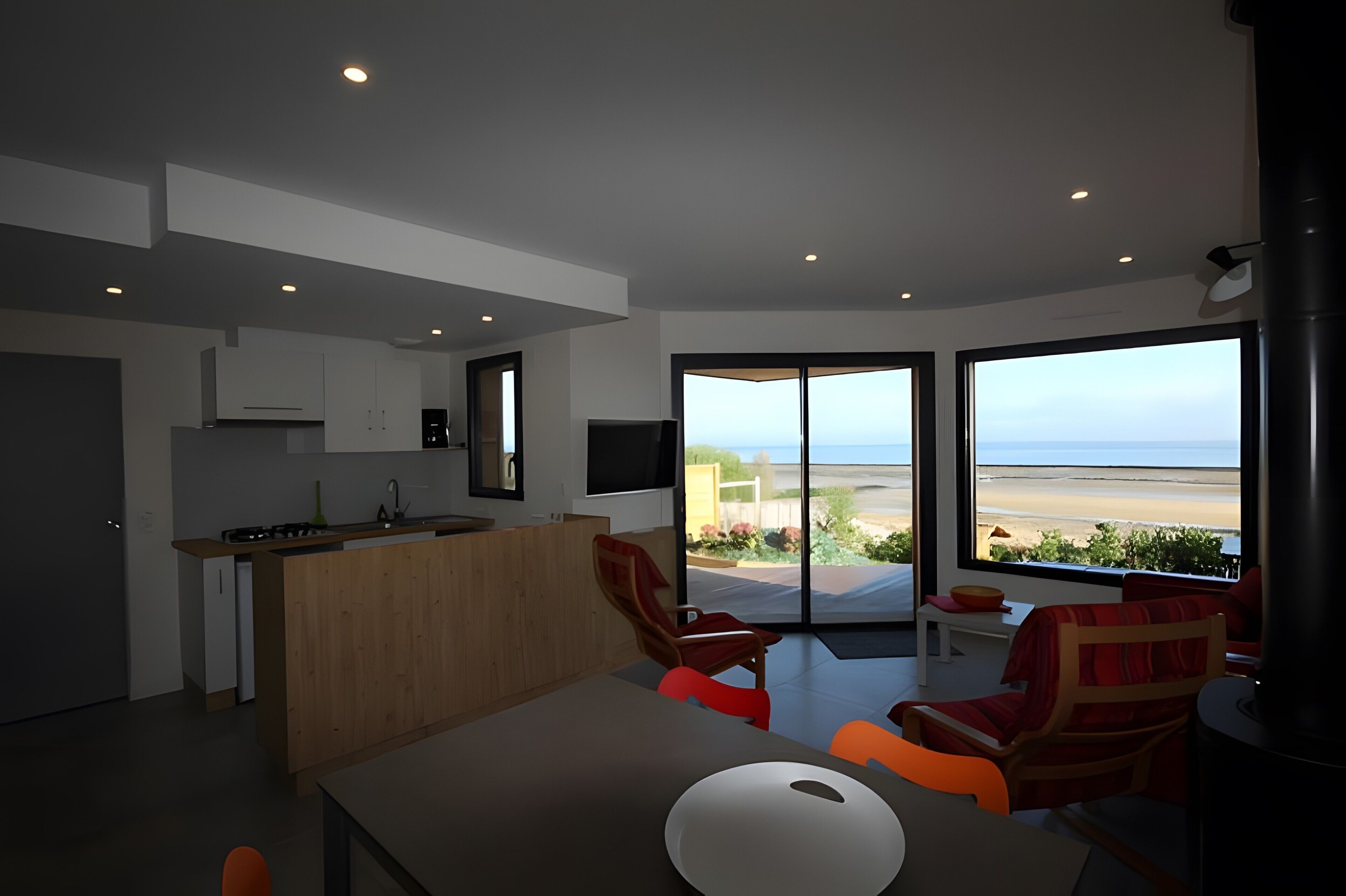 Un intérieur moderne comprenant une cuisine, un coin repas et une grande porte-fenêtre coulissante donnant sur un balcon avec vue sur la plage.