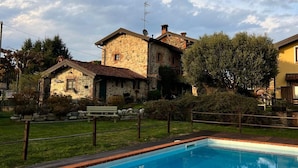 A swimming pool with a stone wall, surrounded by a grassy area and stone buildings. - The Willow Bed & Breakfast - Room Sage (Veruno)