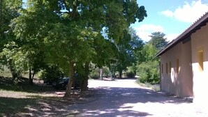 Ein Schotterweg führt zu einem Gebäude mit gelber Fassade und zahlreichen Fenstern. Bäume säumen den Weg, und ein Auto ist links geparkt.