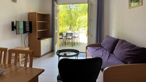 Ein Wohnzimmer mit Fernseher, einer Couch, einem Couchtisch und einem Bücherregal. Eine Tür führt zu einem Außenbereich mit Tisch und Stühlen.