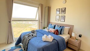 A bedroom with a bed, a nightstand, a ceiling fan, a window with curtains, and a view of the landscape. - Skyline Haven 2BHK 28th Floor + Pool (Gandhinagar)