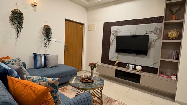 A modern living room with a sofa, coffee table, TV, ceiling fan, and decorative wall lights. - Skyline Haven 2BHK 28th Floor + Pool (Gandhinagar)