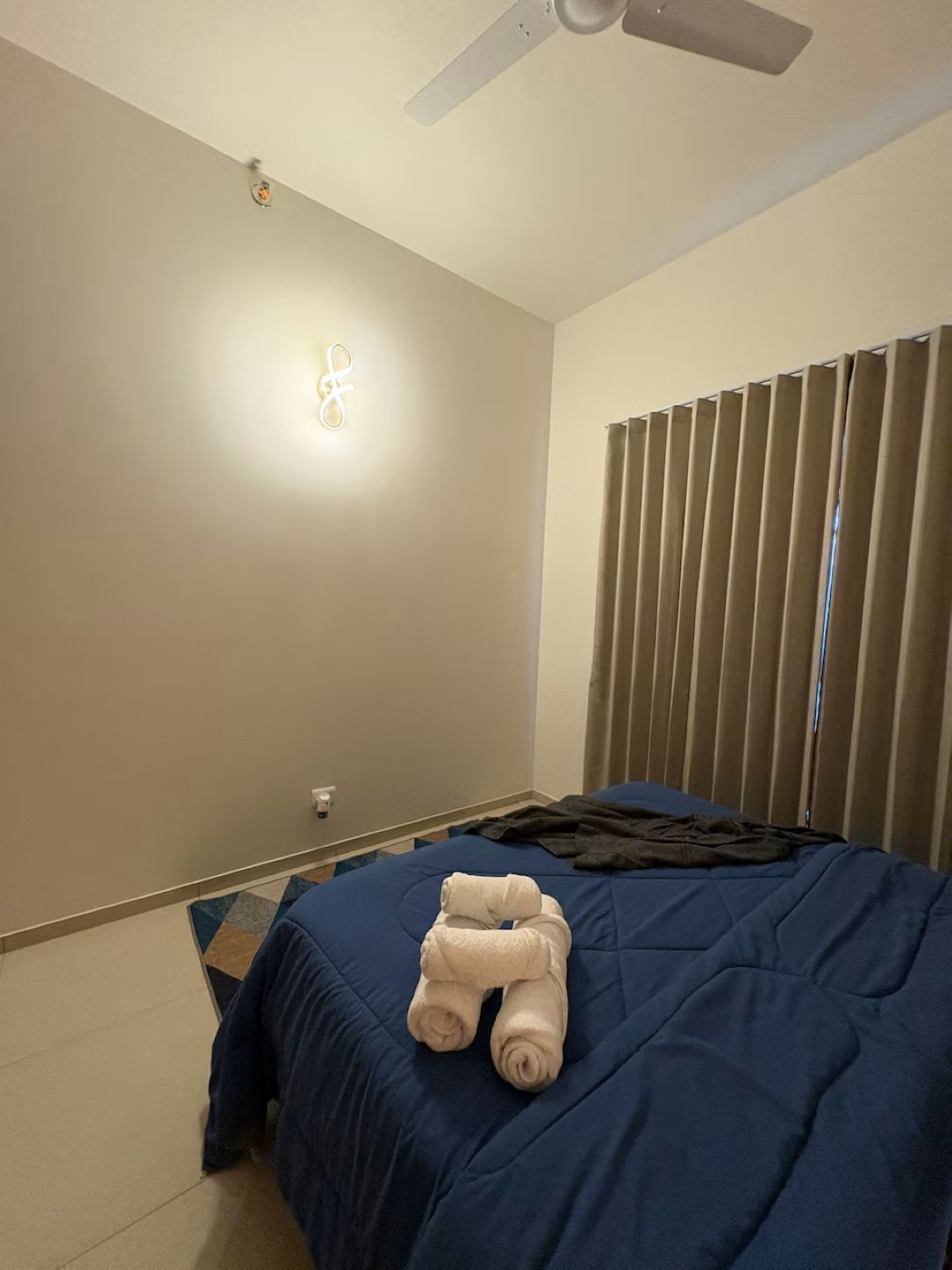 A hotel room with a bed, a ceiling fan, and a wall-mounted light fixture.