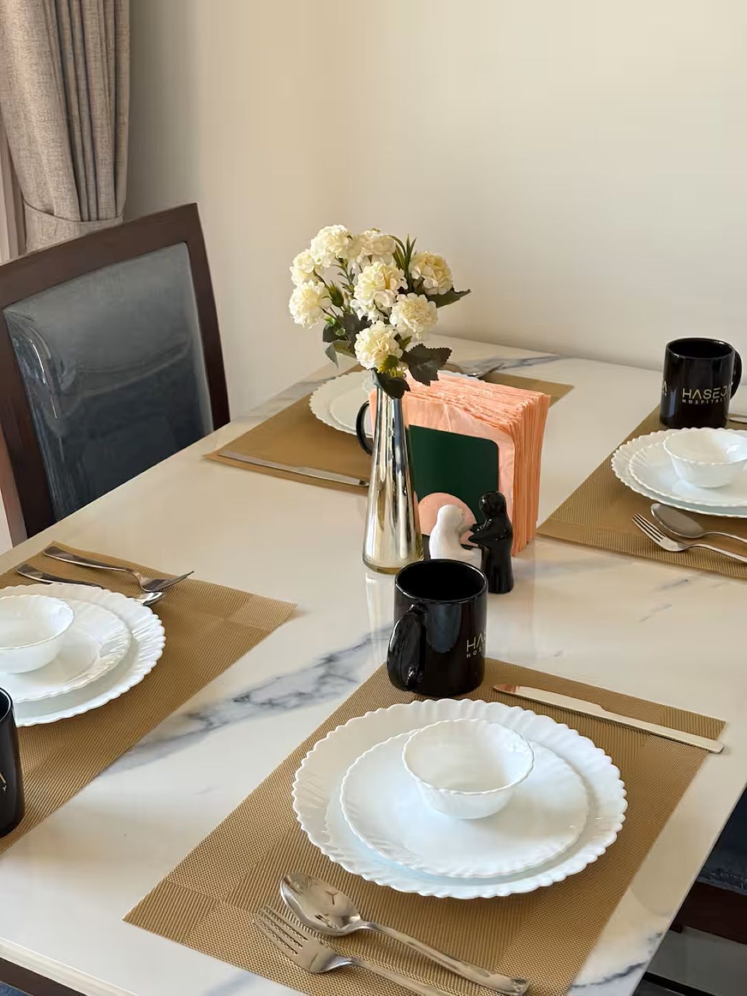 A dining table set with white plates, black mugs, and a vase with white flowers.