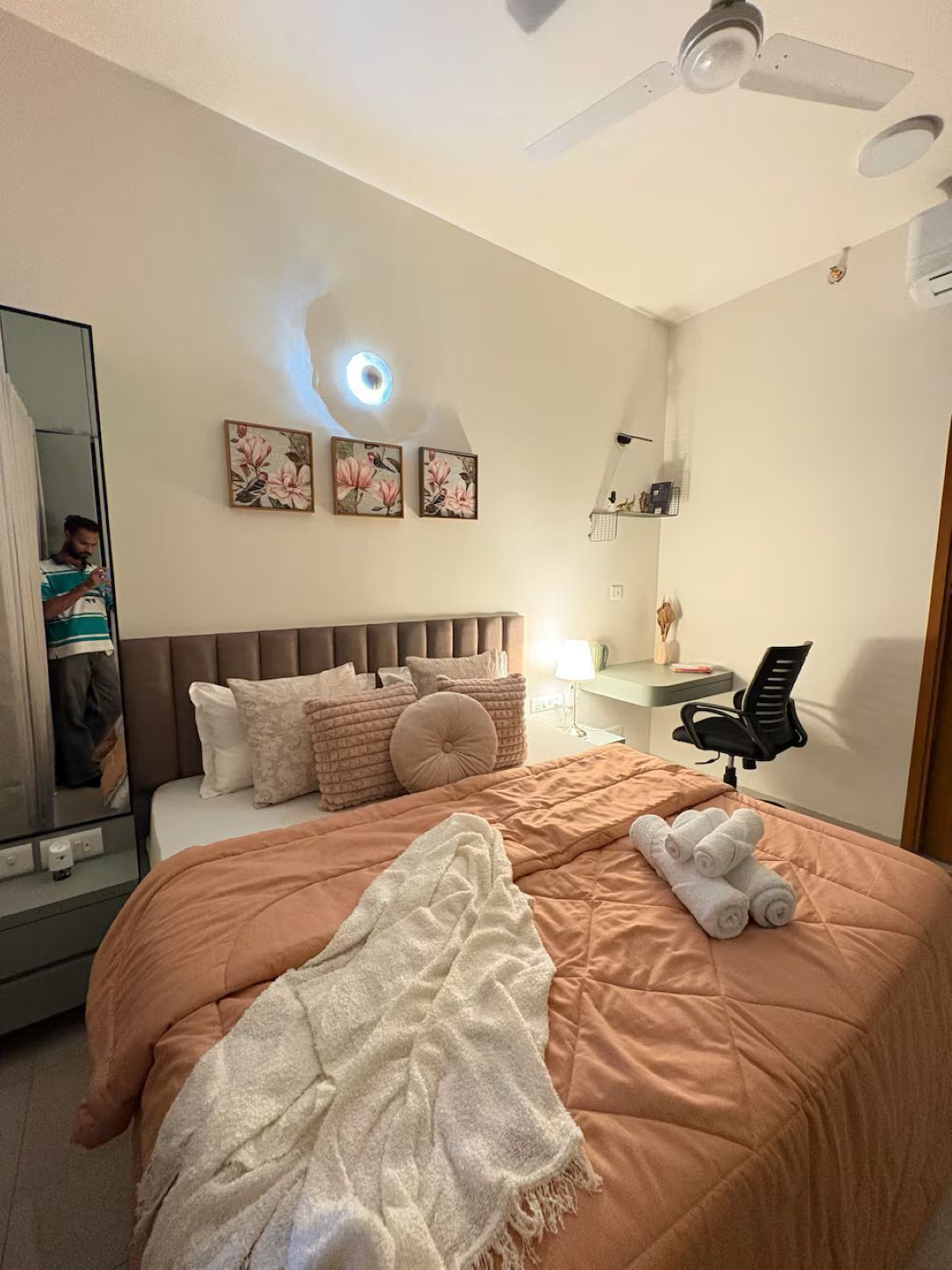 Une chambre à coucher comprenant un lit, un bureau, une chaise, un ventilateur de plafond et des œuvres d’art accrochées au mur.