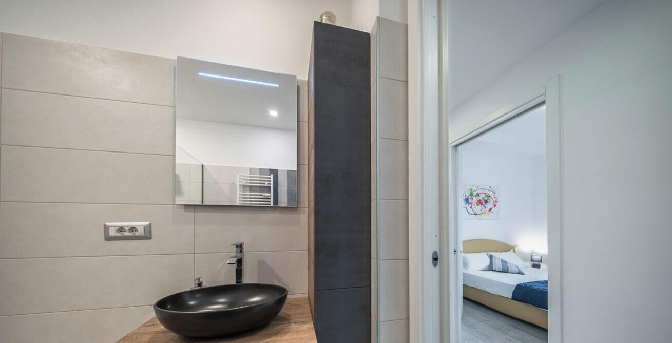 A modern bathroom with a large mirror, a black vessel sink, and a visible shower area.