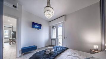 A bedroom with a bed, a bench, a chandelier, a television, and a window.