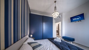 Hotellihuone, jossa on sänky, seinälle asennettu televisio, kattokruunu ja sininen penkki.
