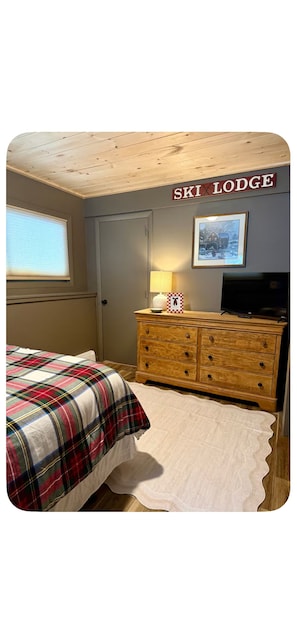 Une chambre à coucher avec une couvre-lit à carreaux, une commode en bois, une télévision et un tableau encadré au mur.