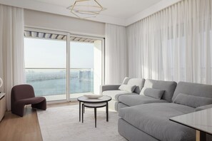 A modern living room with a grey sofa, a small round table, a dark armchair, and a view of a cityscape through large windows. - Elegant 2BR Apartment with Sea View (Dubai)