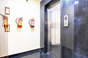 A hallway with a fire extinguisher mounted on the wall, a fire alarm, and an elevator door. - Classic With Balcony Rosedale Reliance Guest House (Barasat)