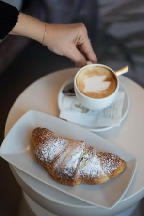 A croissant on a white plate, a cup of coffee with latte art, and a hand reaching for the coffee. - Agorà Luxury Experience (Zafferana Etnea)
