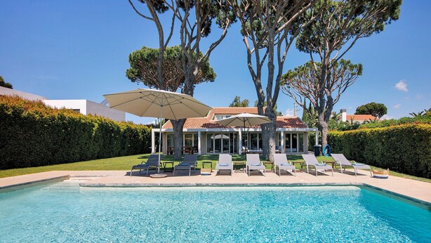 Una piscina con lettini e ombrelloni, circondata da siepi e alberi.