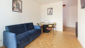 Ein Wohnzimmer mit einem blauen Sofa, einem Esstisch mit Stühlen und zwei gerahmten Bildern an den Wänden.