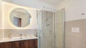 Ein modernes Badezimmer mit einer gläsernen Duschkabine, einem runden Spiegel mit integrierter Beleuchtung und einem Holzwaschtisch.
