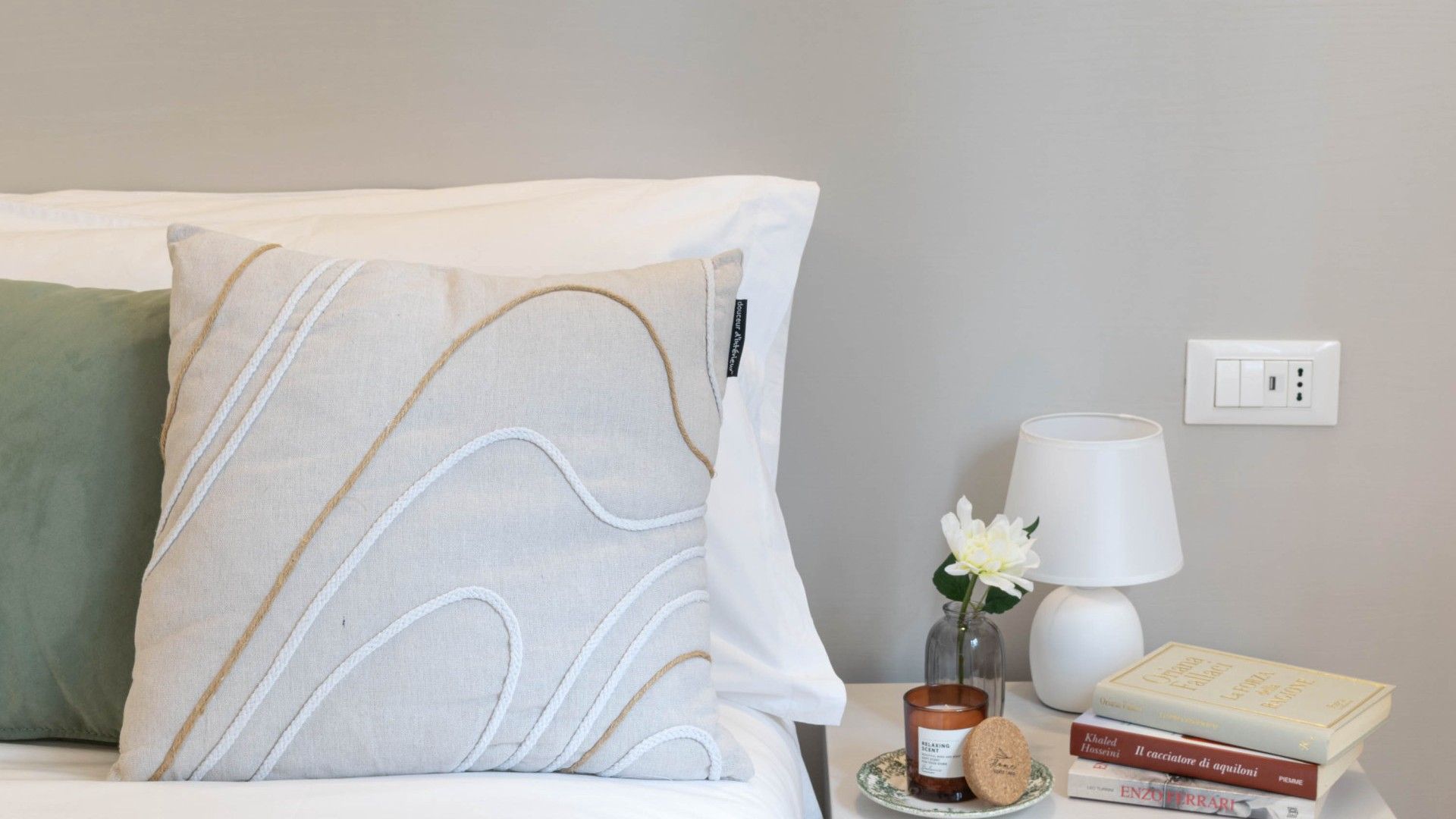 A neatly made bed with a pillow, a bedside table with a lamp, a vase with a flower, and a stack of books.