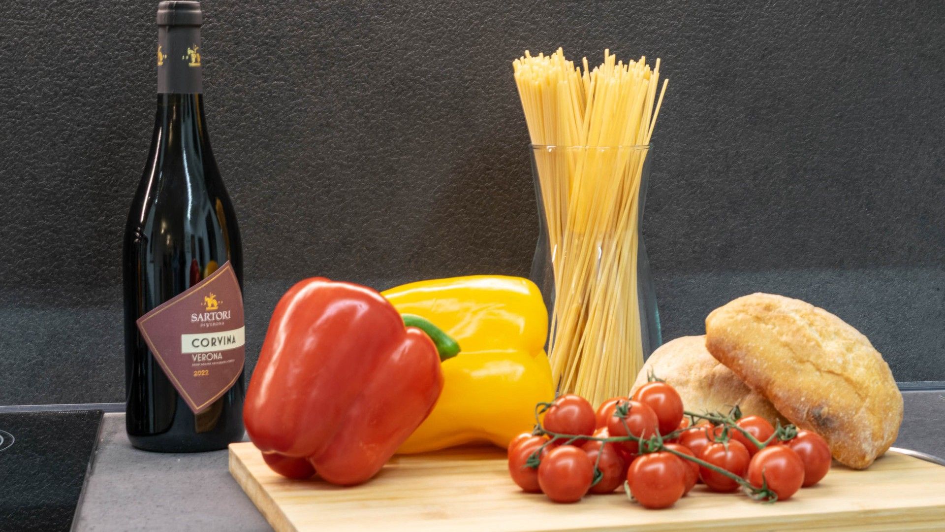 A bottle of wine, pasta, a bell pepper, cherry tomatoes, and bread on a cutting board.