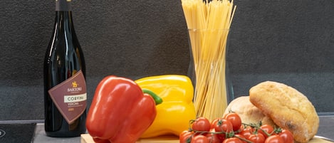 A bottle of wine, pasta, a bell pepper, cherry tomatoes, and bread on a cutting board.