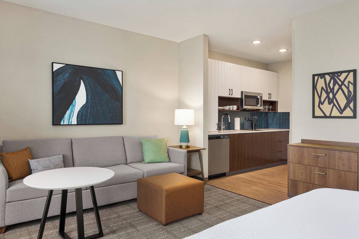Ein modernes Hotelzimmer mit einer Couch, Hocker, einem kleinen Tisch, einer Lampe, einem Bett, einer Küchenzeile und Kunstwerken an den Wänden.