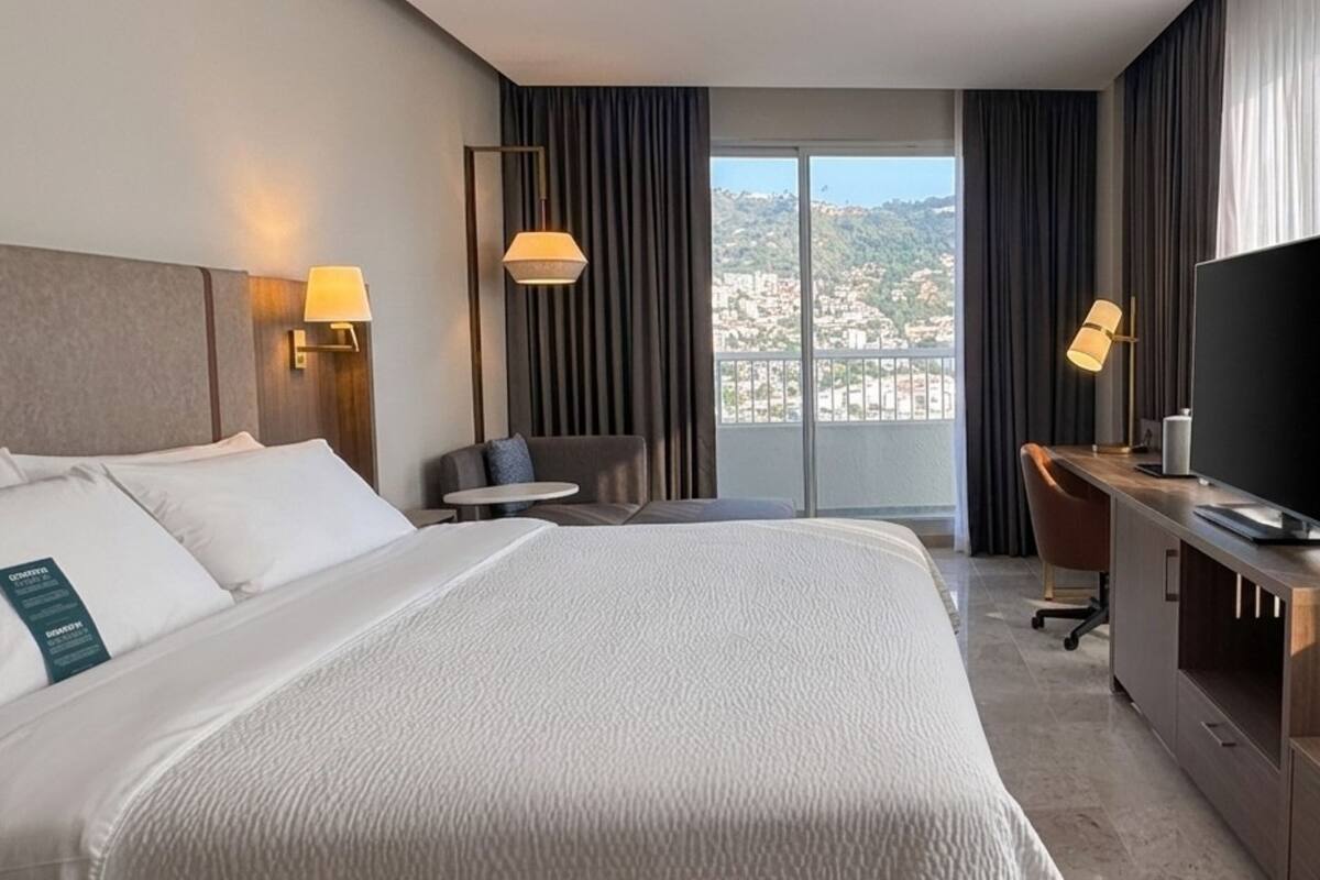 A hotel room with a bed, a desk, a television, and a view of a cityscape.