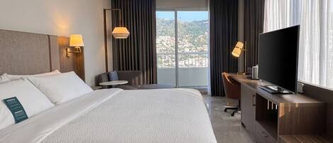 A hotel room with a bed, a desk, a television, and a view of a cityscape.