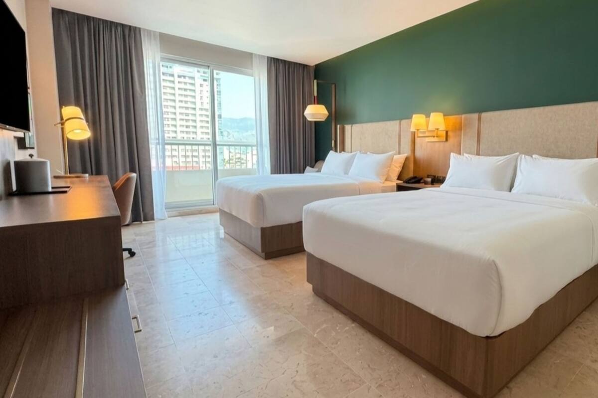 A hotel room with two beds, a desk, a chair, a lamp, and a view of a building through the window.