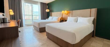 A hotel room with two beds, a desk, a chair, a lamp, and a view of a building through the window.