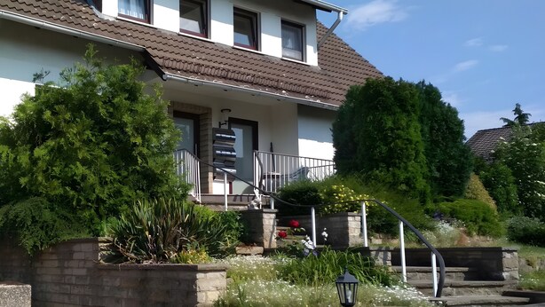 A two-story house with a brown tiled roof, a front porch with a staircase, and a garden with various plants and flowers. - Apartment "For 4 Guests, 75 m²" with Shared Garden and Wi-Fi (Heeßen)