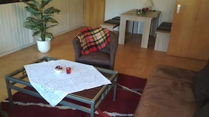 Ein Wohnzimmer mit einem Glastisch, einem braunen Ledersofa, einer rot-schwarz karierten Decke auf einem braunen Lederstuhl, einer Topfpflanze und einem kleinen Tisch mit Lampe und Laptop.