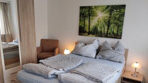 A bedroom with a bed, a chair, a nightstand, a lamp, and a painting of a forest. - Apartment "For 4 Guests, 75 m²" with Shared Garden and Wi-Fi (Heeßen)