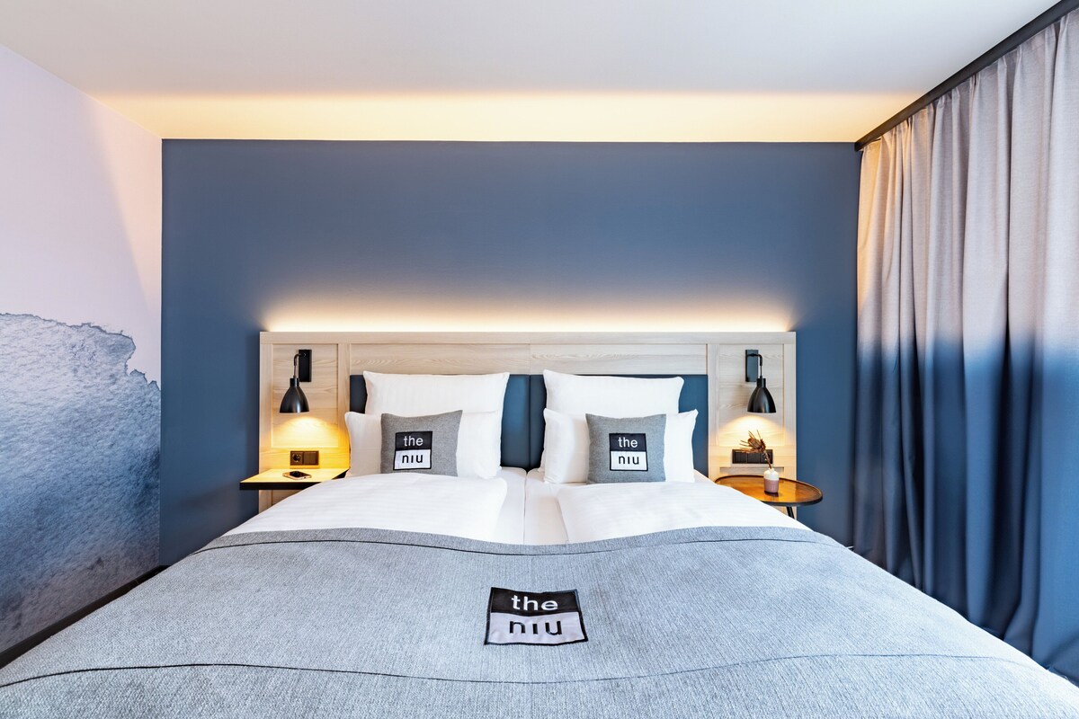 Cama arrumada com um cobertor cinza, duas almofadas com o logotipo 'the niu' e abajures laterais.