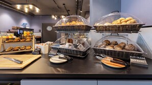 A breakfast buffet with various baked goods and bread, including dark buns, in a modern setting with a counter and display shelves.