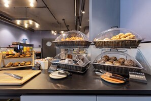A breakfast buffet with various baked goods and bread, including dark buns, in a modern setting with a counter and display shelves. - Holiday Inn - The Niu, Quay Hamburg Harburg By IHG (Hamburg)