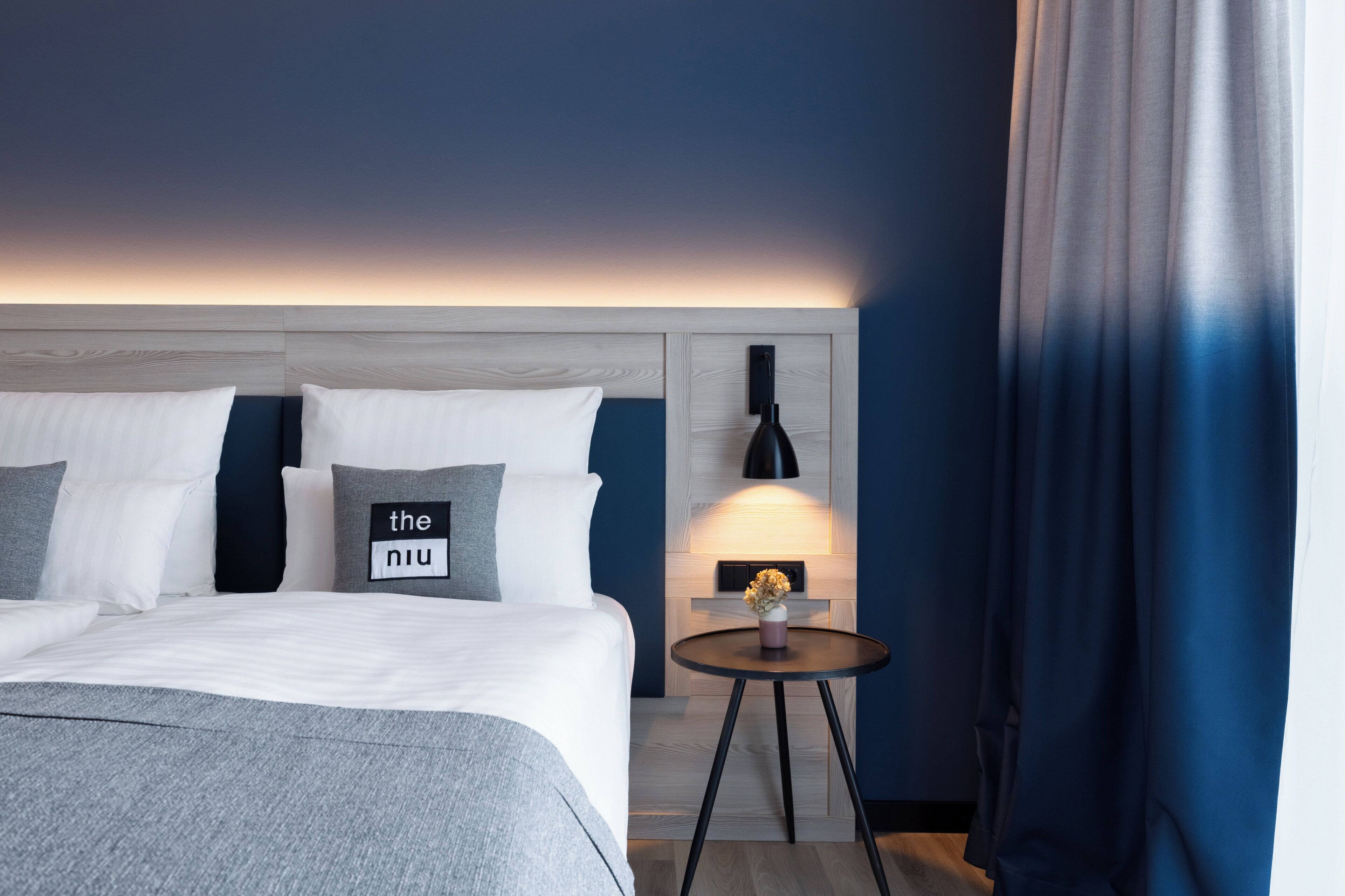 Een hotelkamer met een bed, een nachtkastje met een lamp en een bedieningspaneel aan de muur.