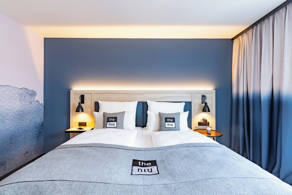 Un lit bien fait, avec une couverture grise, deux oreillers ornés du logo « the niu » et des lampes de chevet.