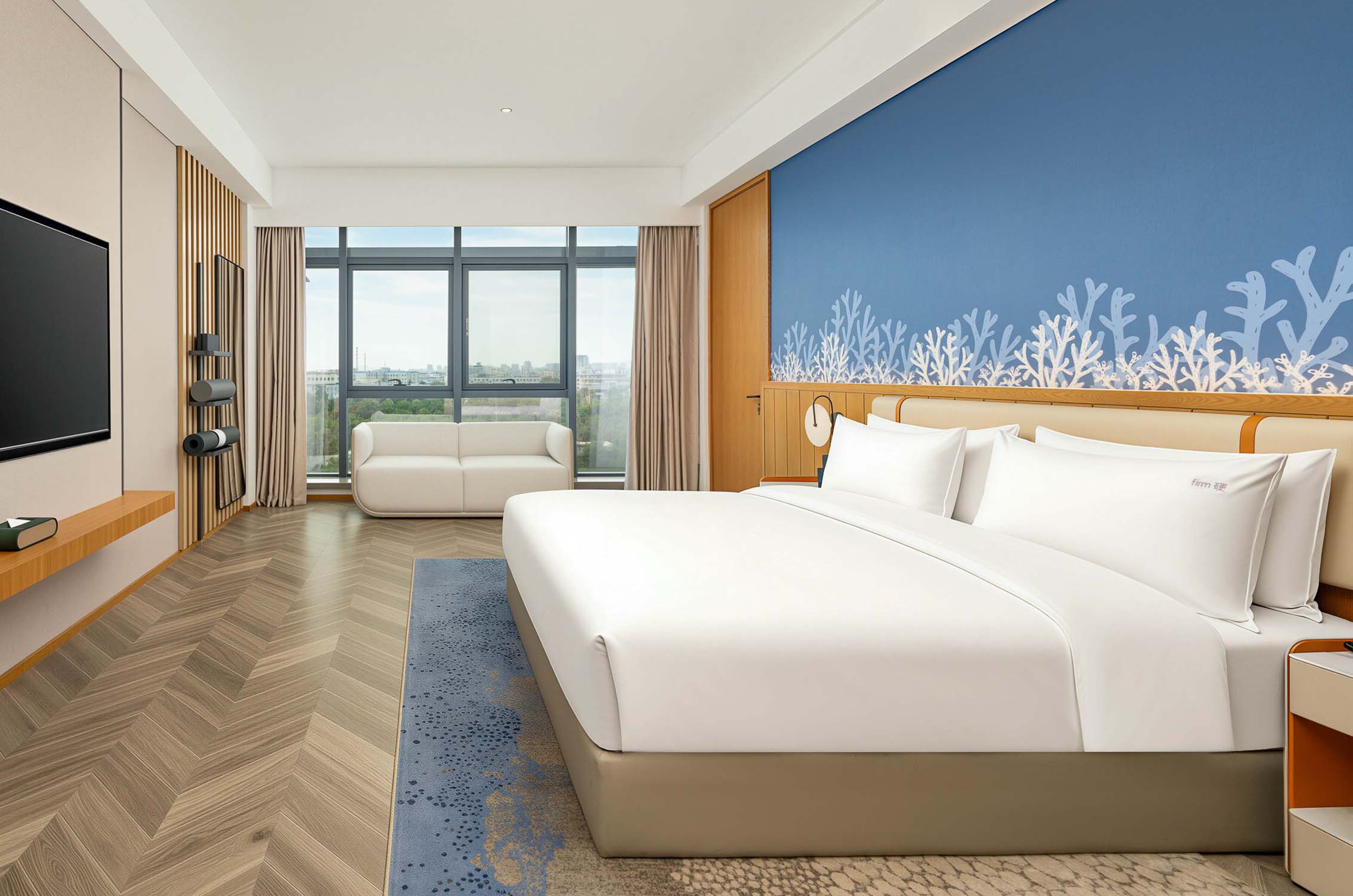 Habitación de hotel con una cama grande, televisión, zona de estar con sofá y vistas a la ciudad a través de las ventanas.
